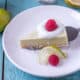 Overhead view of a slice of a paleo key lime pie garnished with coconut cream and a raspberry being placed on a white plate with a silver spatula next to a slice of lime and a raspberry.