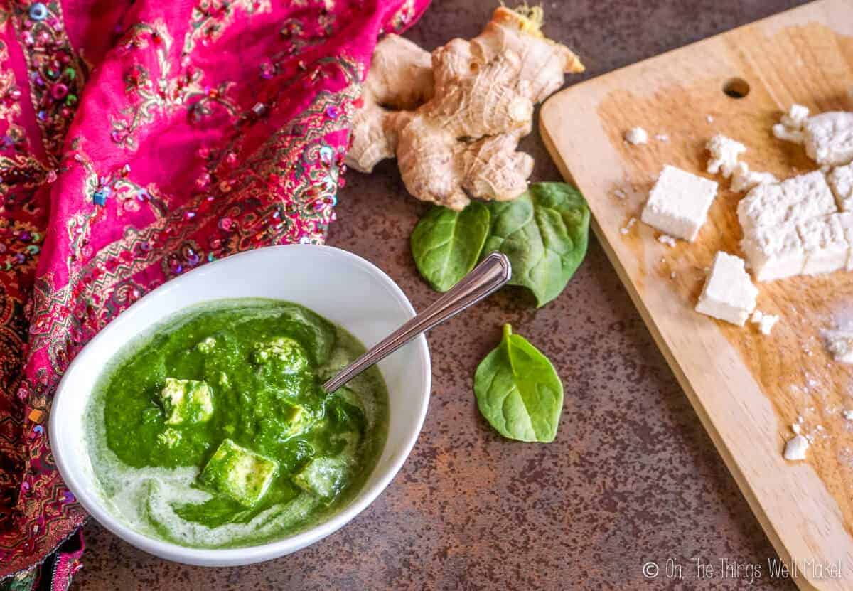 overhead view of palak paneer next to some cut paneer, some fresh spinach leaves, and a ginger root.