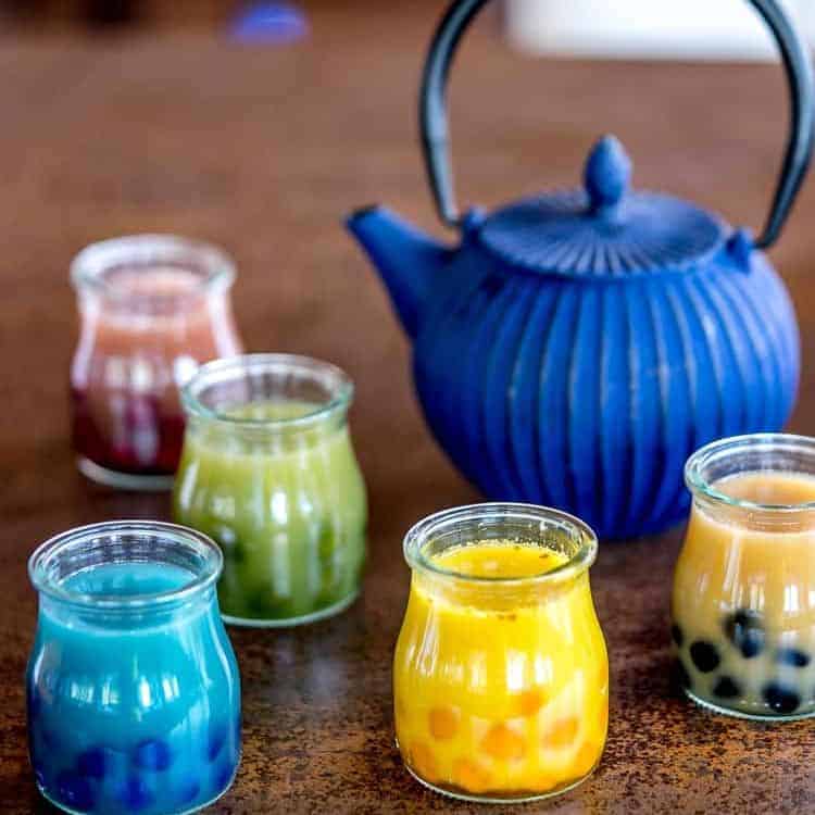 Glasses of colorful tea with colorful boba tapioca pearls in it with a blue cast iron tea pot in the background.