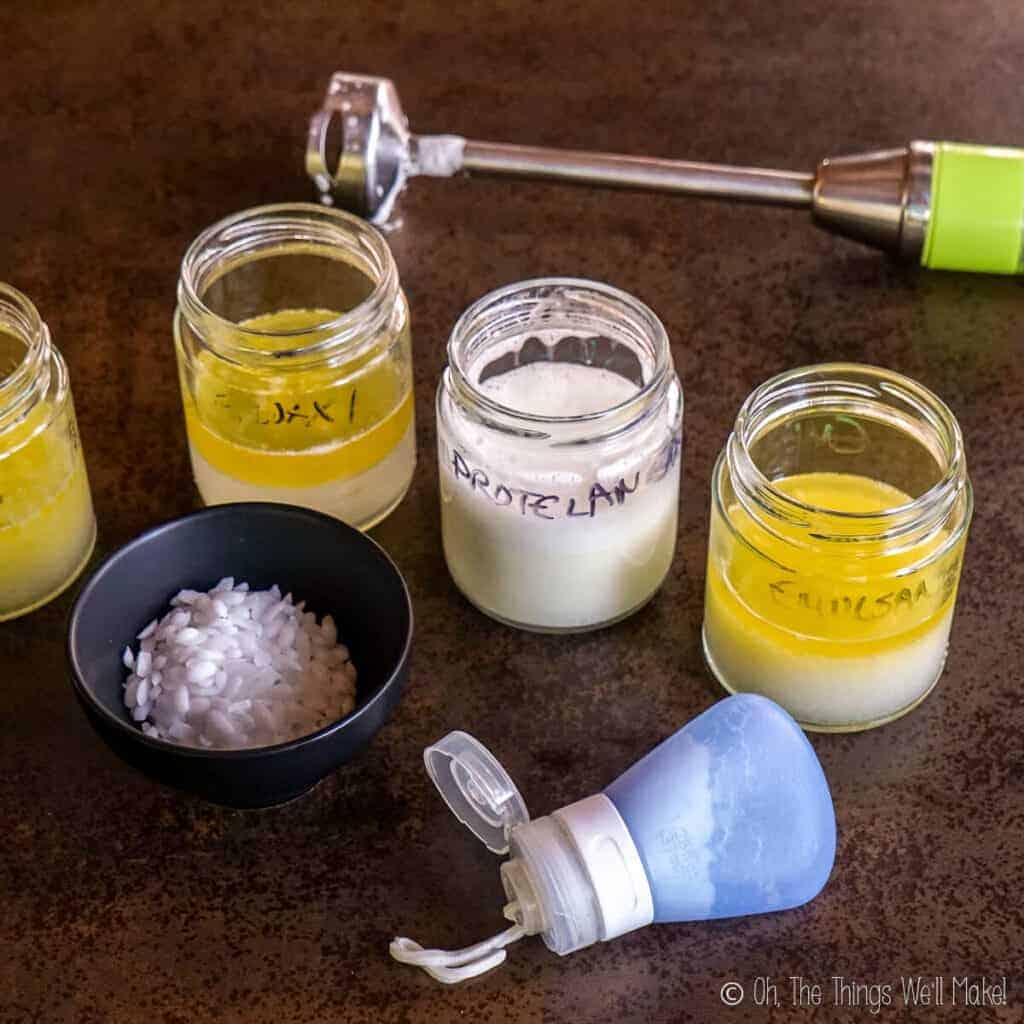Several jars of lotion being made- showing different states of being emulsified. Behind them is an immersion blender. In front of them in a finished lotion and a bowl of emulsifying wax.