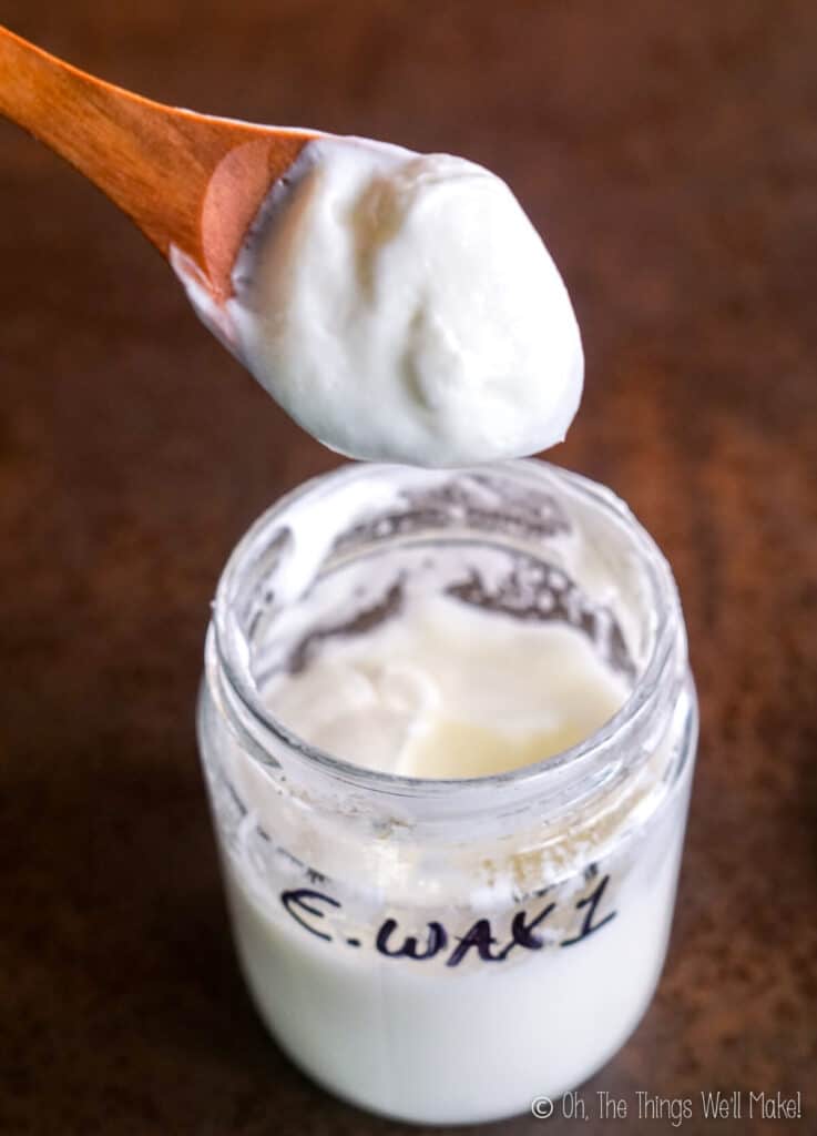 A wooden spoon filled with a lotion made with emulsifying wax #1 held over a jar of the lotion