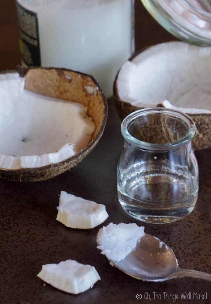 A small jar of melted coconut oil in front of a jar of solid coconut oil- both next to an open coconut. 