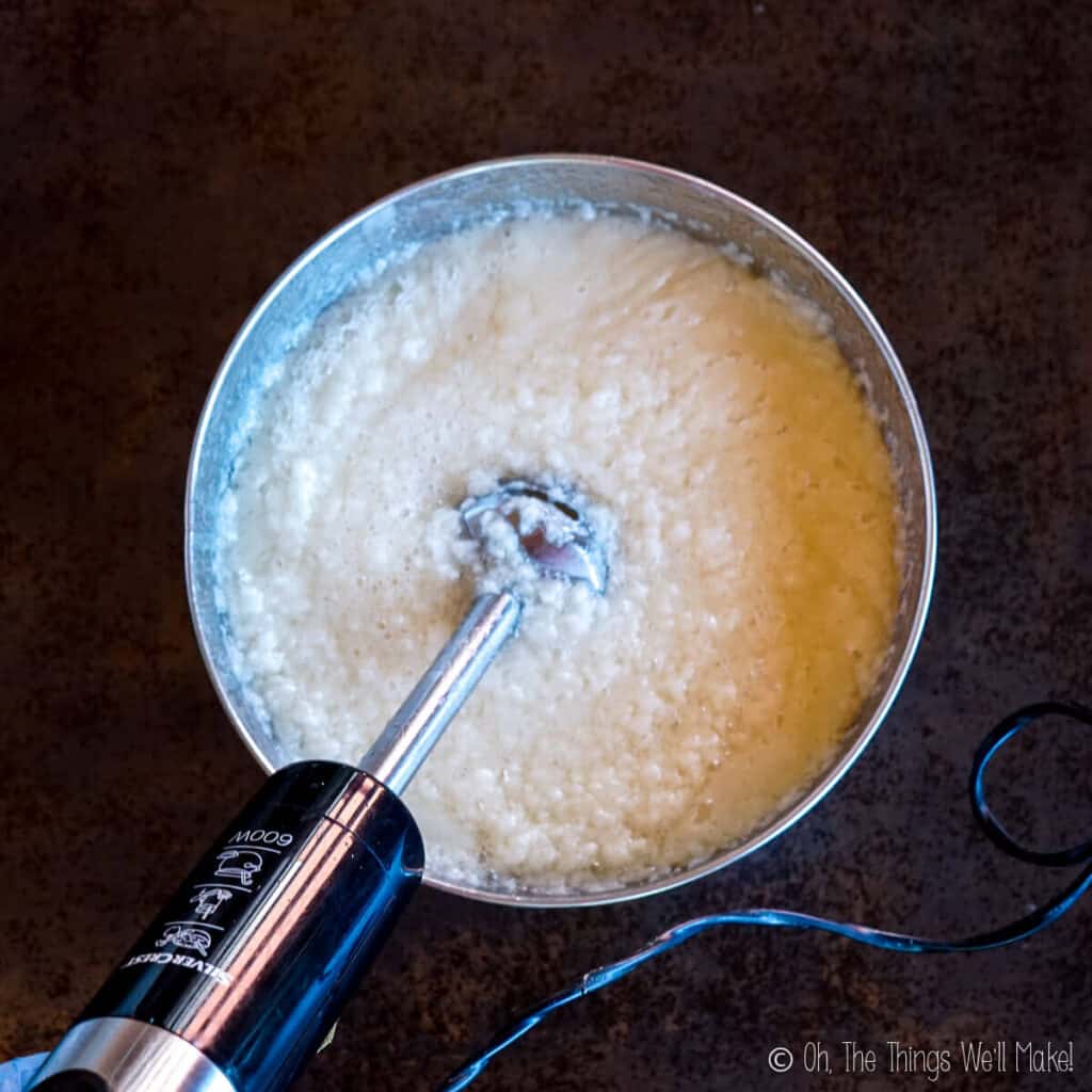 A grainy looking soap mixture in a stainless steel bowl with an immersion blender in it. 