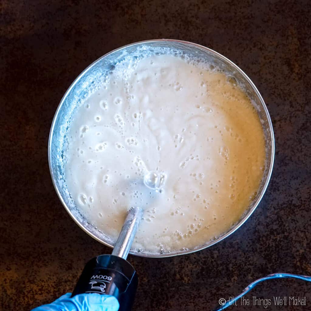 A thick creamy soap mixture in a stainless steel bowl with an immersion blender in it.