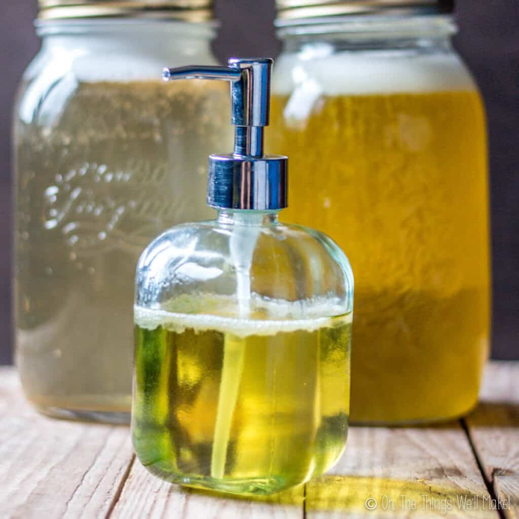 A bottle of liquid soap in a dispenser in front of two jars of different liquid soaps