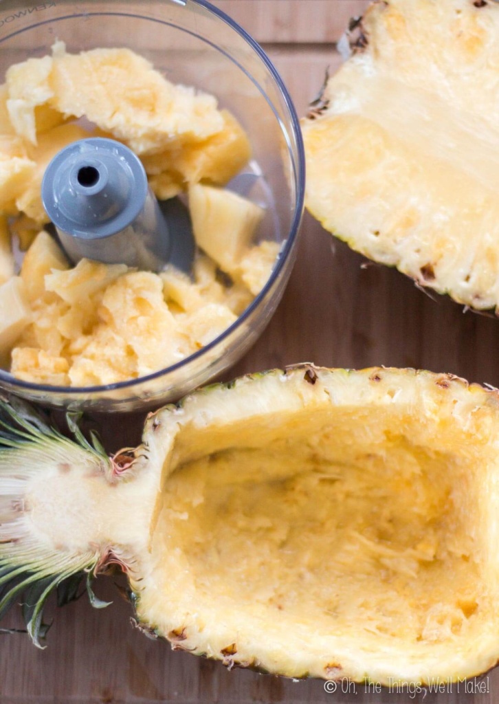 Frozen pineapple in a food processor next to a hollow pineapple rind