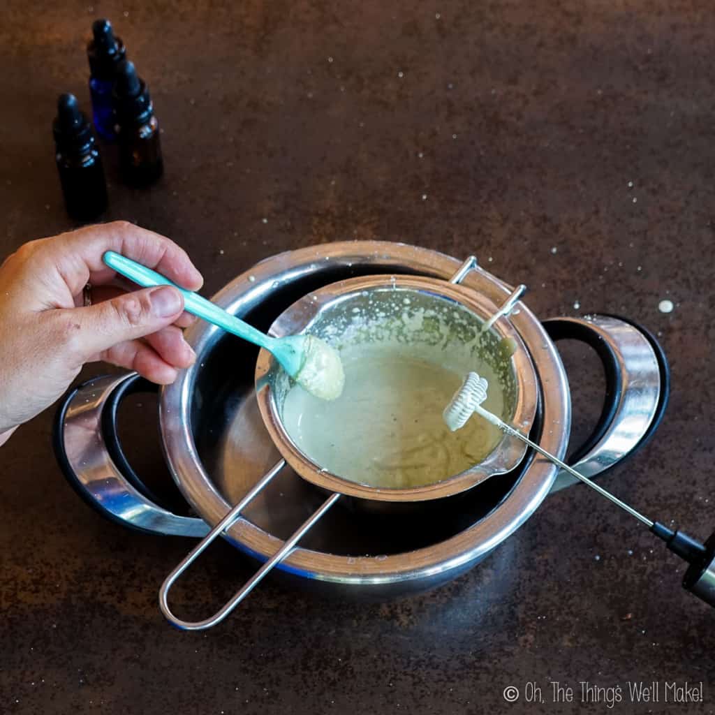 lifting up some of the deodorant mixture on a spoon after having blended it with the milk frother shown resting on the double boiler.