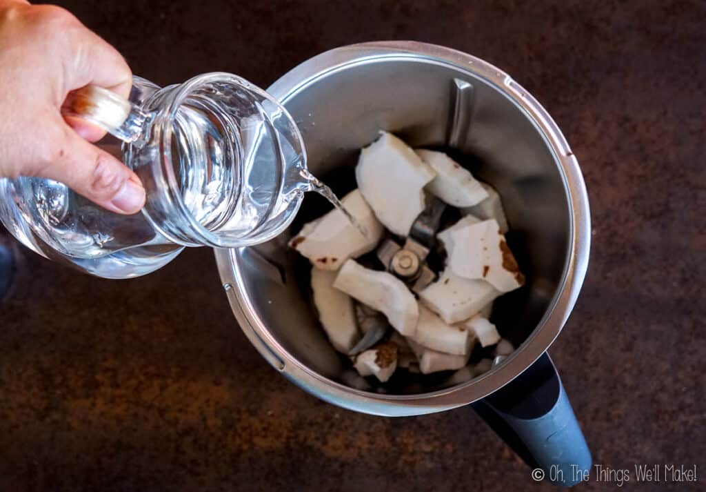 Pouring water from a pitcher into a food processor with coconut pieces