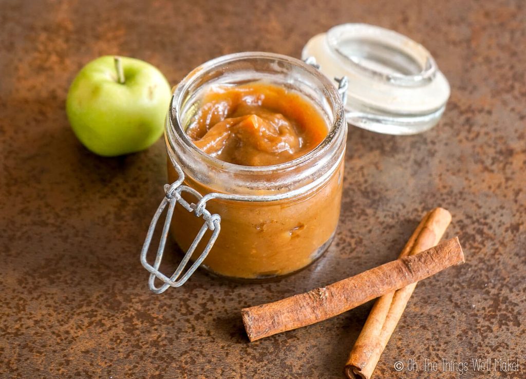 Homemade spiced apple butter in an open glass jar with a green apple and cinnamon sticks.