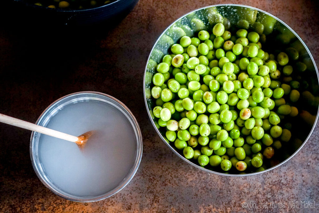 A bowl of green olives next to a bowl ith lye solutions