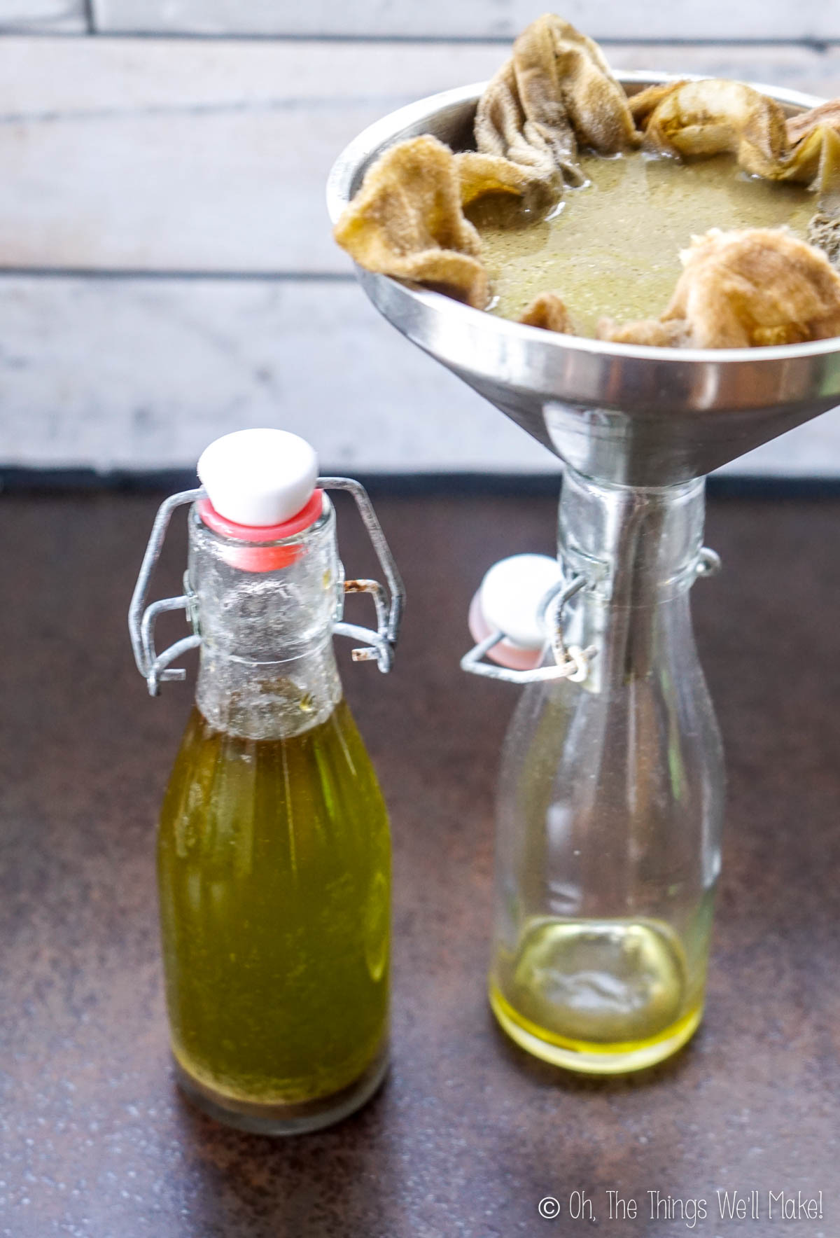 One full bottle of olive oil and one with oil being filtered into it through a cloth filled funnel filled with an olive mixture