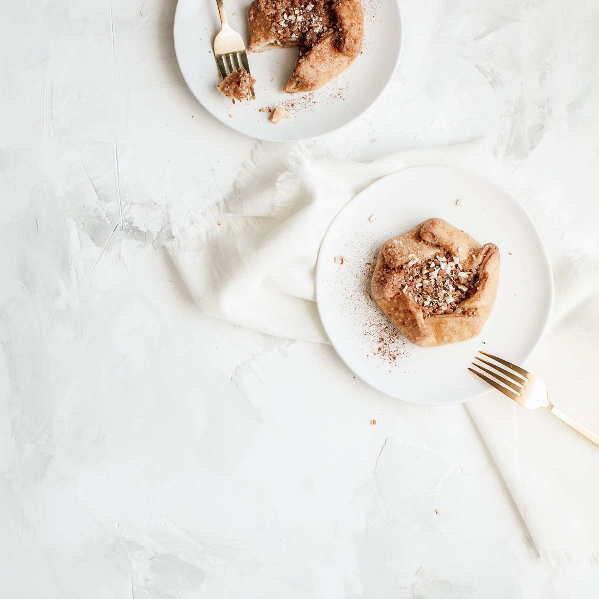 Two small pastries with powdered topping on white plates, one partially eaten, placed on a textured white surface with gold forks and a white napkin.