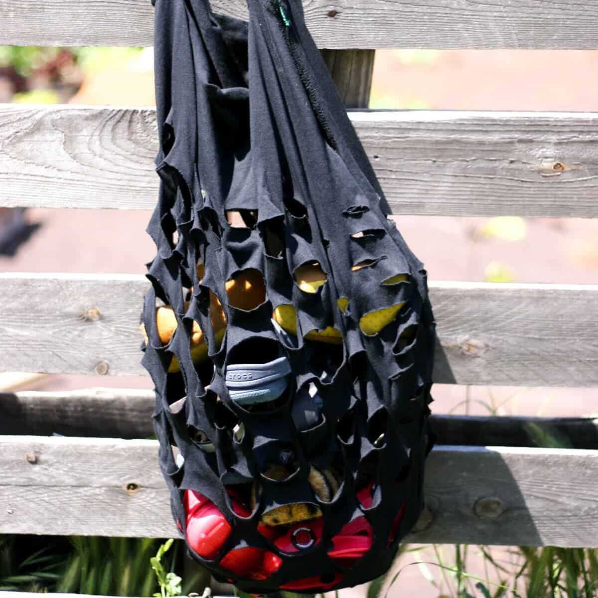 A handmade t-shirt black produce style bag with slits cut into it, filled with beach items hanging on a wood fence.