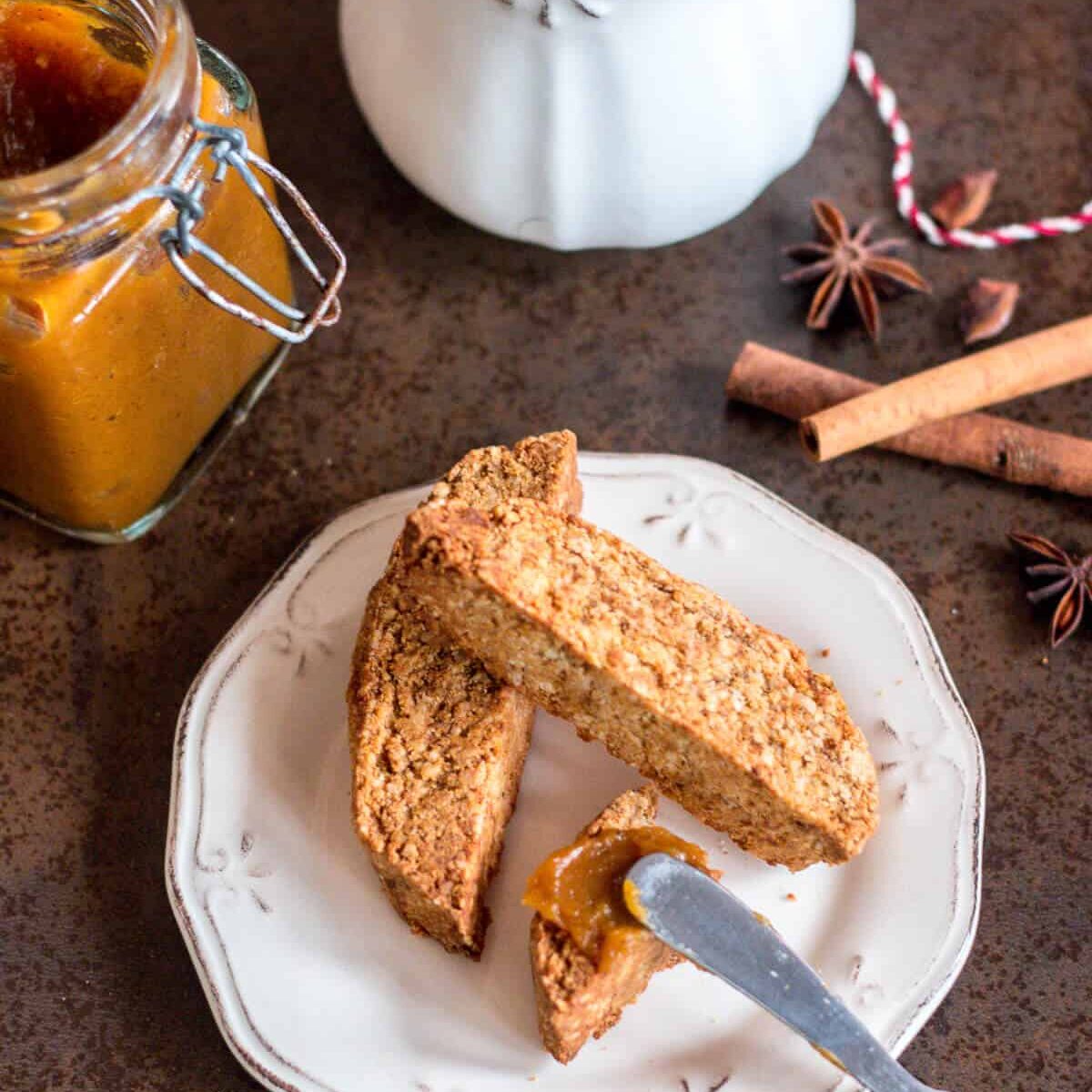 Top view of three pumpkin spice biscotti on a plate with a knife spreading spiced pumpkin butter over one piece. Beside the plate are an open jar of pumpkin butter, a white ceramic jug, and a couple pieces of cinnamon sticks and star anise seeds.