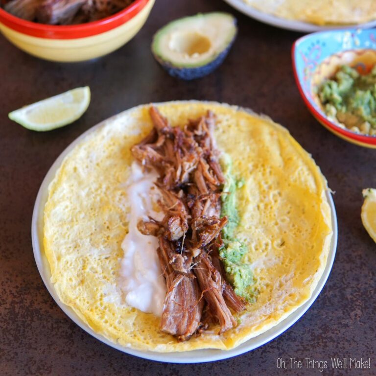 A tortilla filled with savory carnitas, sour cream, and guacamole is displayed on a plate. There is a half avocado, a lime wedge, and other food items in the background.