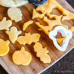 Cheese slices cut into Halloween shapes like ghosts, bats, and pumpkins on a wooden board with a cookie cutter.