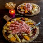 A charcuterie board with a meat and cheese hand design on a wooden board, accompanied by a purple candle and another board with assorted snacks in the background.