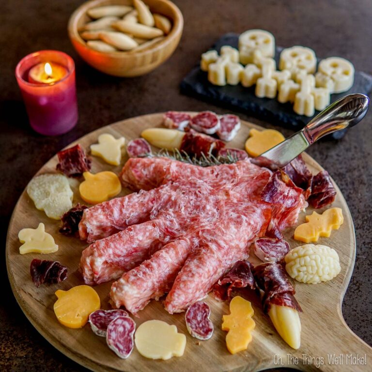 A charcuterie board features a Halloween hand-shaped cheese ball centerpiece, surrounded by various cheeses and meats, with a lit candle and breadsticks in the background.