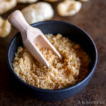 A black bowl filled with pork rind crumbs, with a wooden scoop resting inside.