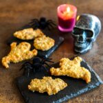 Halloween-themed chicken nuggets in ghost and bat shapes on slate plates, with decorative spiders, a lit pink candle, and a silver skull in the background.