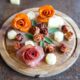 A Valentine's Dy charcuterie board a wooden board with arranged meat and cheese shaped into roses, surrounded by small leaf garnishes.