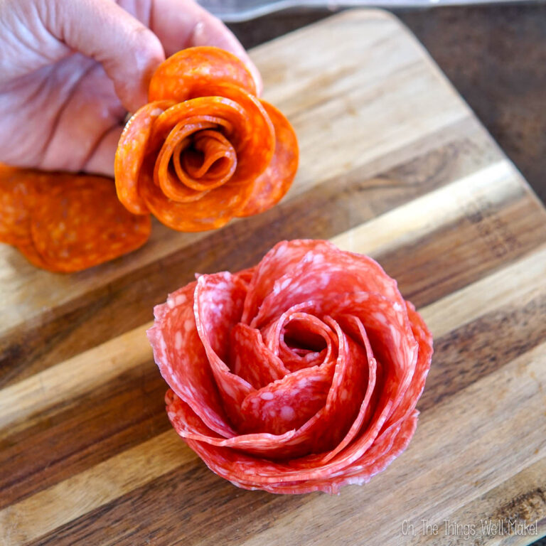 Hand holding a flower made of pepperoni slices above a wooden cutting board with salami rose.