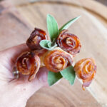 A hand holding a bouquet of bacon roses with green sage leaves, set against a wooden background.