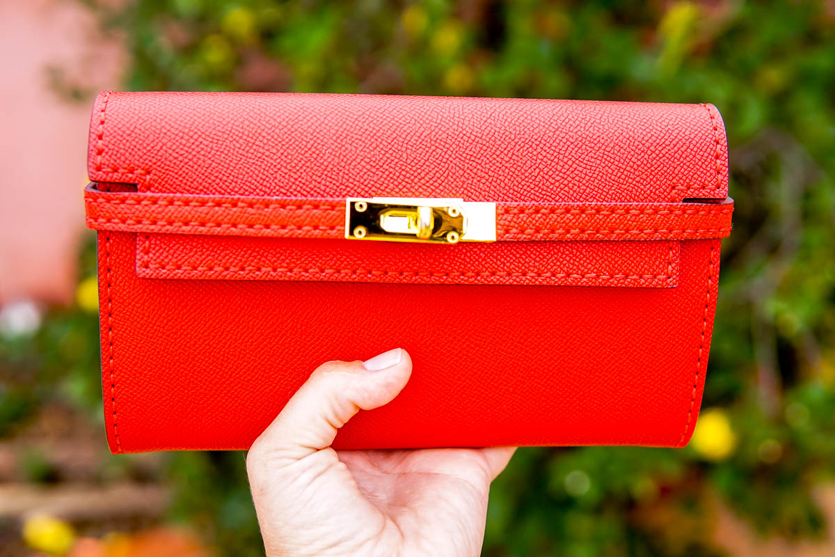 A hand holding up a vibrant red Kelly clutch purse with a gold clasp.