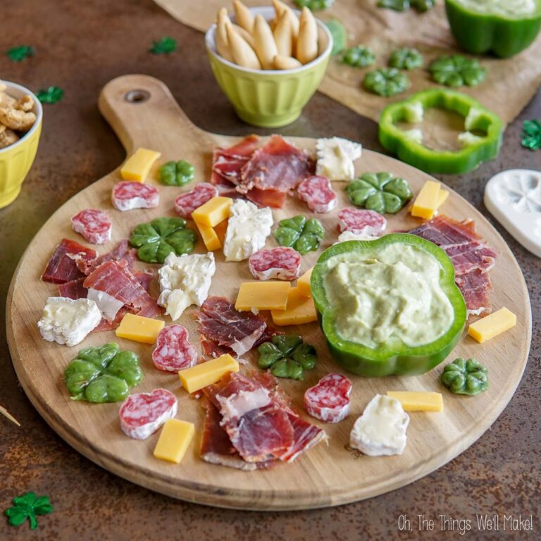 A wooden board with assorted cheeses, cured meats, and green bell pepper filled with dip. Green clover-shaped crackers and dip bowls are nearby. Small shamrock decorations are scattered around.