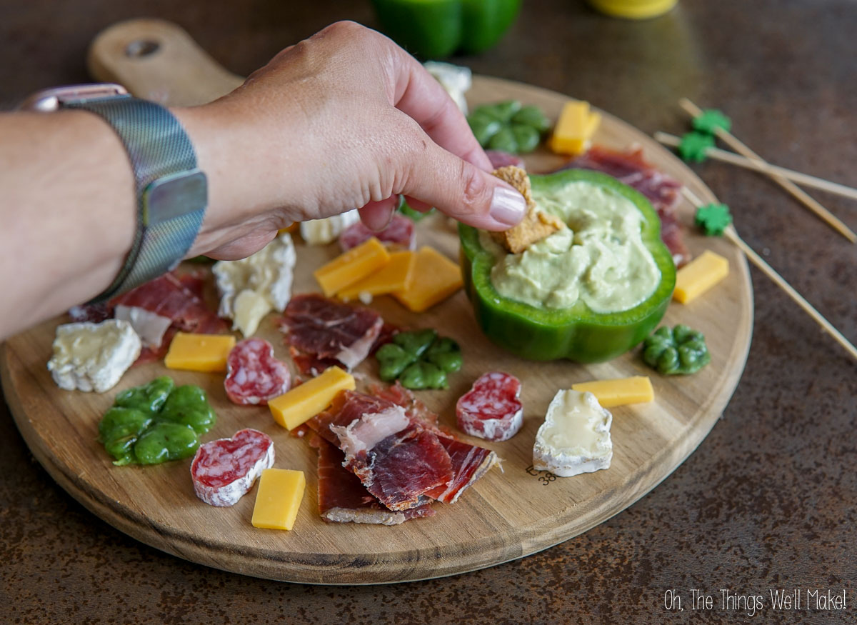 A hand dips a pork rind into an avocado cream dip served in a hollowed green pepper on a wooden board, creating the perfect St. Patrick's Day charcuterie board with assorted cheese and cured meats.