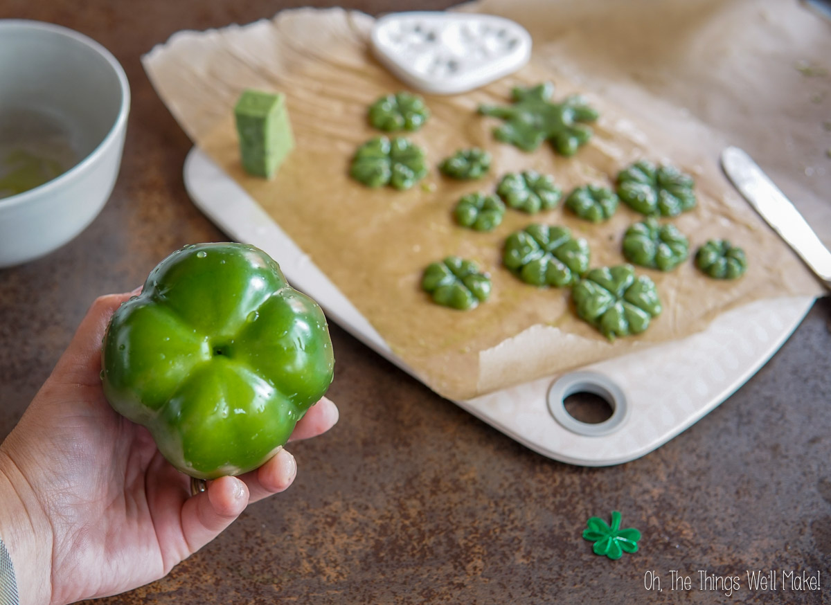 Person holding a green bell pepper next to a cutting board with bell shamrock-shaped green cheeses on parchment paper.