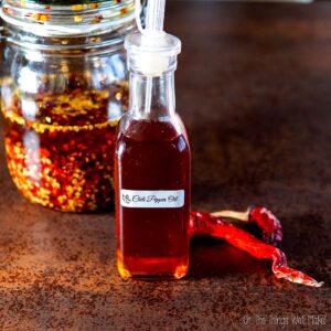 Bottle labeled "Chili Pepper Oil" with infusion of chili flakes and oil; dried red chilies beside the bottle; glass jar with chili flakes and oil in the background.