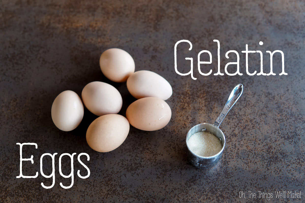 Six brown eggs are arranged on a dark surface next to a metal measuring cup filled with powdered gelatin. The words "Eggs" and "Gelatin" are labeled near each item.