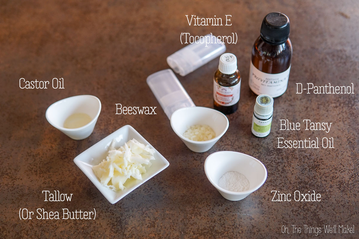 Various skincare ingredients in bowls and bottles, including castor oil, beeswax, tallow, vitamin E, D-panthenol, blue tansy essential oil, and zinc oxide, displayed on a table.