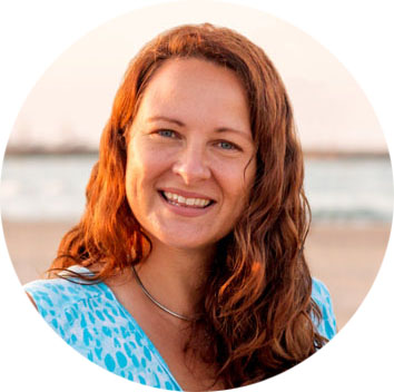 A woman with long, wavy brown hair smiles at the camera while standing on a beach. She is wearing a blue patterned top and a silver necklace. The background shows sand and water.