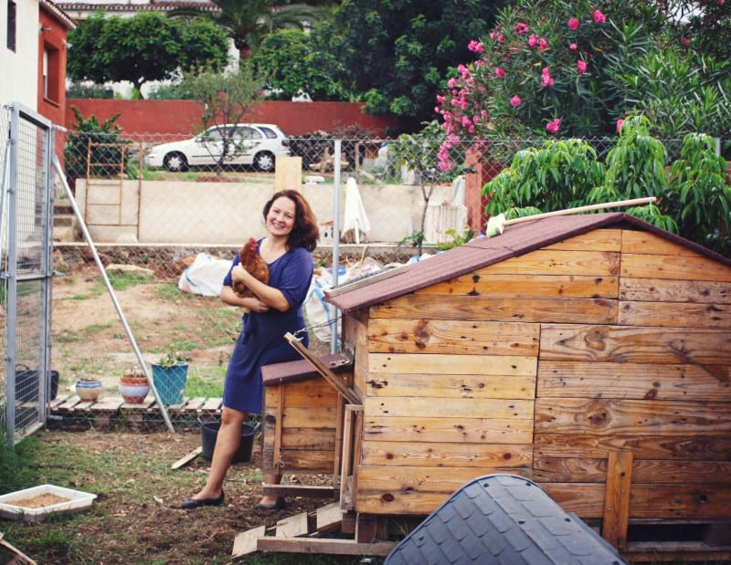 Dr. Tracy Ariza in a blue dress stands beside a wooden chicken coop in a garden, holding a chicken and smiling at the camera.