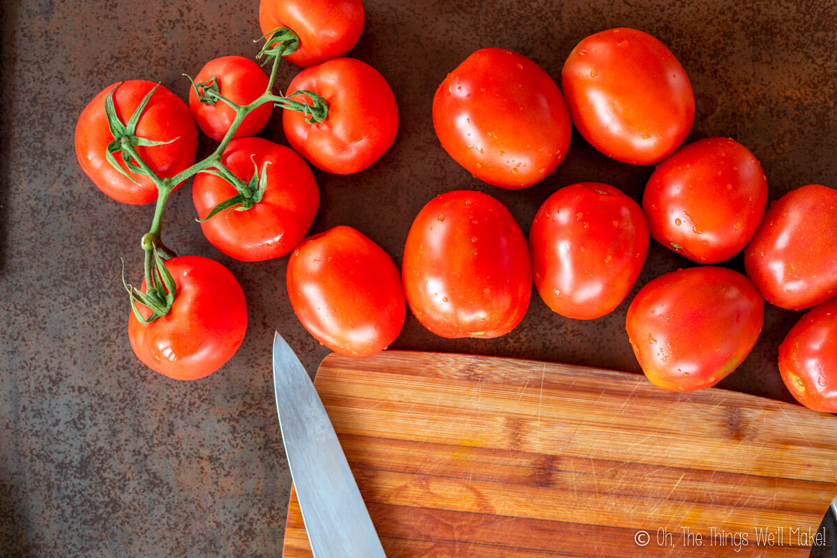 A cluster of vine tomatoes and several loose Roma tomatoes rest on a brown surface next to a knife, a wooden cutting board, and a jar of tomato paste.