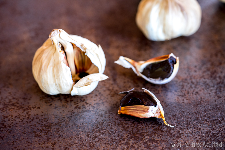 A head of black garlic with two peeled black garlic cloves and one unpeeled clove rests on a brown textured surface.