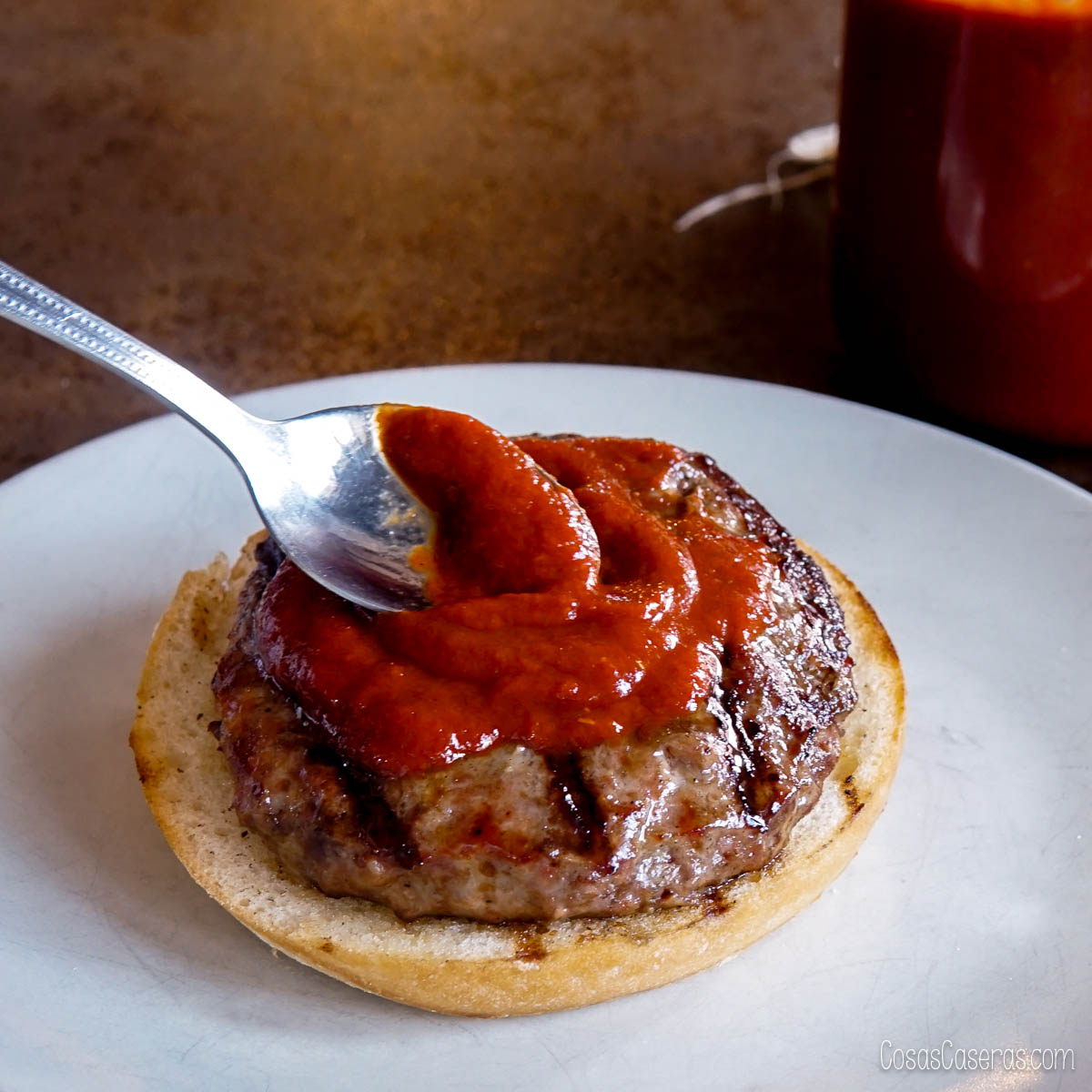 A spoon spreading homemade ketchup on a grilled beef patty placed on the bottom half of a hamburger bun, with a jar of homemade ketchup in the background.