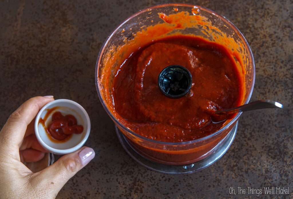 A hand holds a small dish with ketchup next to a food processor bowl filled with homemade ketchup and a spoon inside.