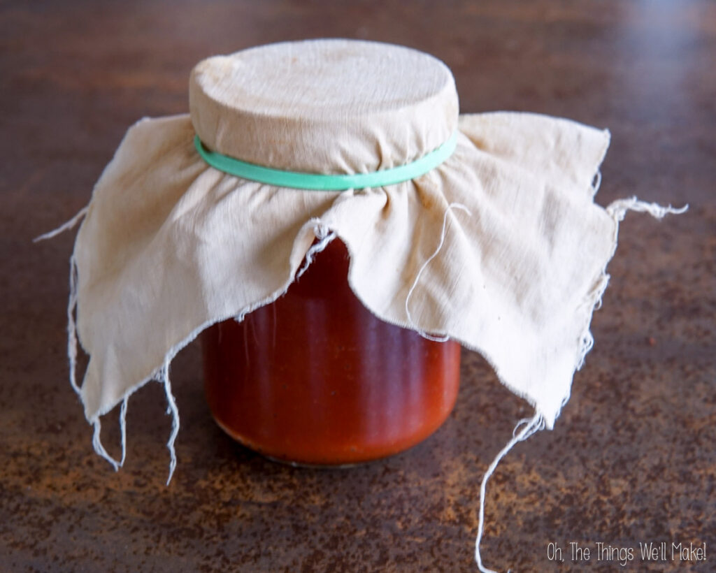 A glass jar filled with homemade ketchup, covered with a cloth secured by a rubber band, sitting on a brown textured surface.