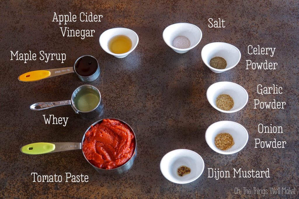 Ingredients for homemade ketchup recipe arranged on a table: tomato paste, whey, maple syrup, apple cider vinegar, dijon mustard, salt, celery powder, garlic powder, and onion powder.