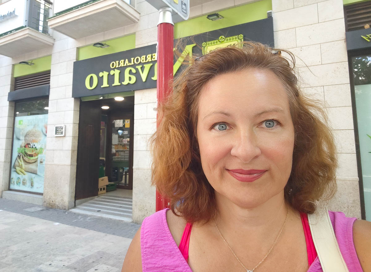 A woman with shoulder-length curly hair stands outside a store called "Herbolario Navarro" on a sunny day, smiling at the camera.