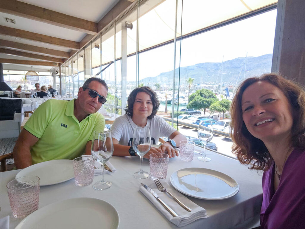 Three people sit at a set dining table in a bright restaurant with large windows overlooking a marina and mountains.