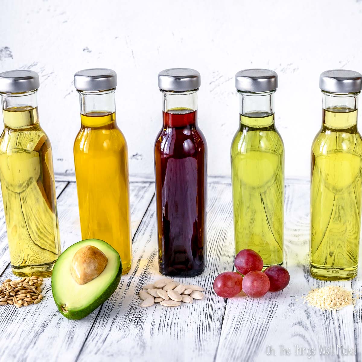 Five glass bottles of various carrier oils stand in a row, each with its source in front: flaxseeds, avocado, pumpkin seeds, grapes, and sesame seeds on a white wooden surface.