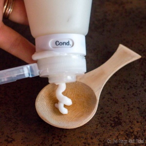 A hand squeezes customized hair conditioner from a bottle labeled "Cond." onto a wooden spoon.