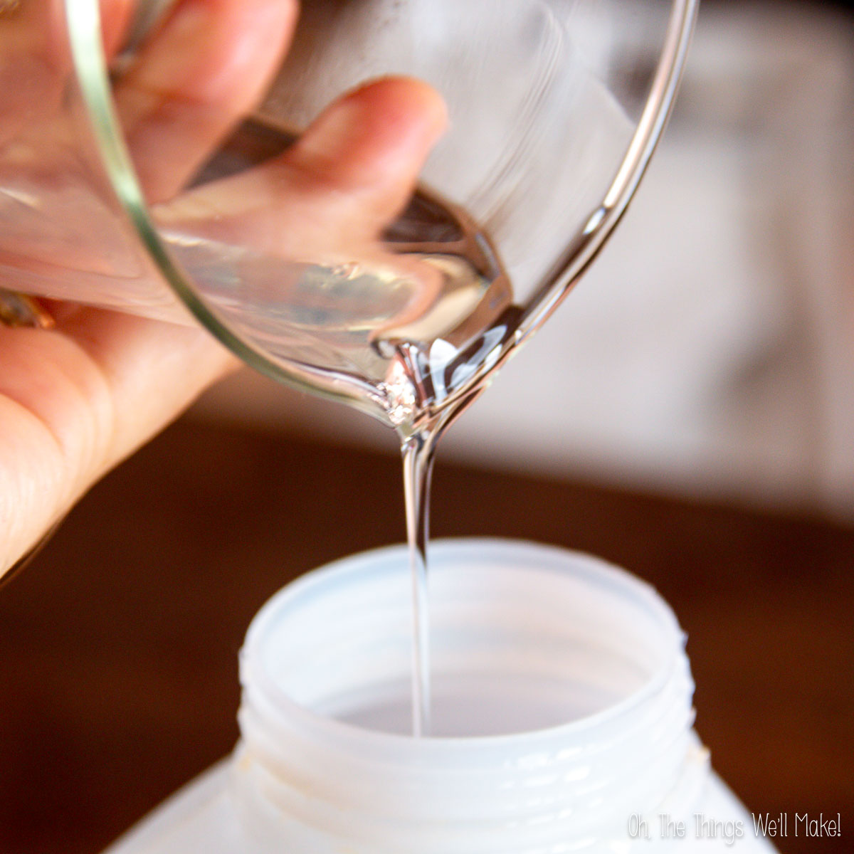 A clear, DIY liquid laundry detergent, is being poured from a glass container into a white plastic bottle.