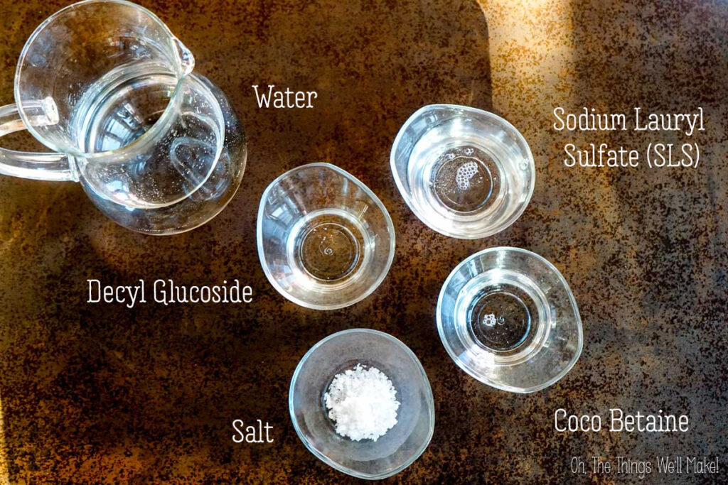 Five clear glass containers on a brown surface, each labeled with their contents&mdash;water, sodium lauryl sulfate (SLS), decyl glucoside, salt, and coco betaine&mdash;perfect for creating your own DIY liquid laundry detergent.