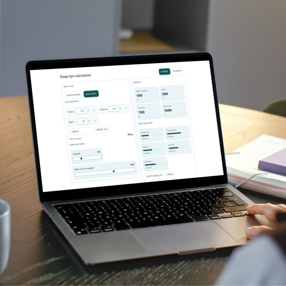 A person uses a laptop displaying a lye calculator interface with input fields and calculated values on a wooden desk.