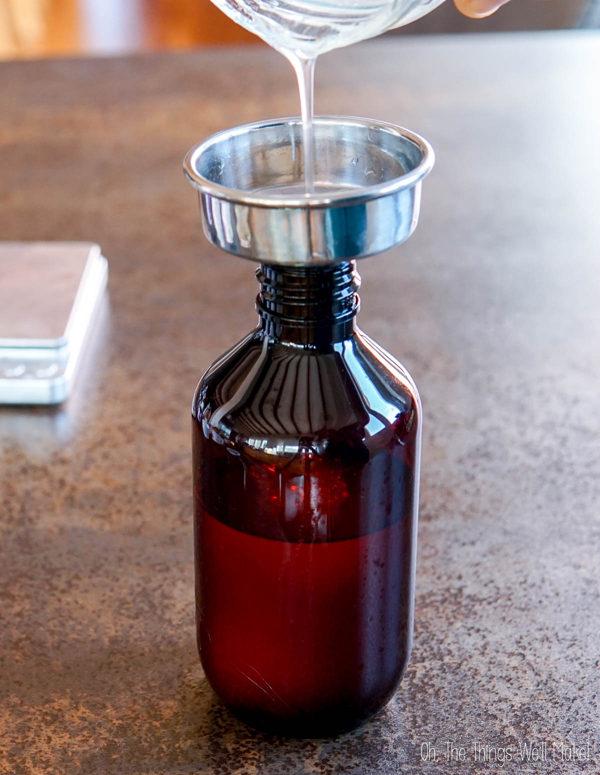 Homemade daily use shampoo is being poured through a metal funnel into a brown glass bottle on a countertop.